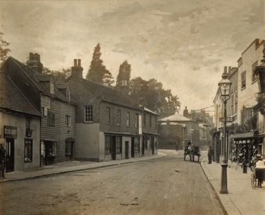 Looking south along the High Street from a position level with the top of Old Bridge Street around 1900. The policeman in the far distance is standing on what would now be the middle of the roundabout at the end of Kingston Bridge. The sign of the "White Hart" can be seen above the head of the cart driver. The shop that was selling cigars is now The Machaan Restaurant.